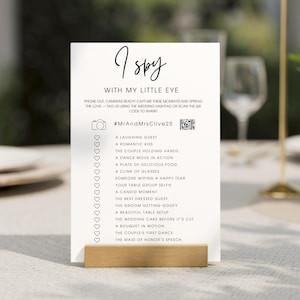 Könnte beinhalten: Eine weiße Karte mit dem Text "I Spy" und einer Liste von Hochzeitsaufforderungen. Die Karte wird in einem Holzständer gehalten. Der Text enthält einen Hashtag und einen QR-Code zum Teilen von Fotos.