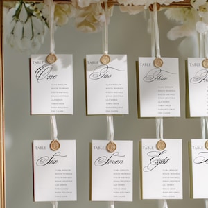 May include: Elegant wedding table seating chart with nine cards, each labeled with a number and names. The cards are white with gold accents and are suspended from a gold frame, adorned with white flowers.
