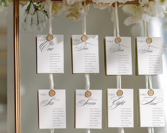 May include: Elegant wedding table seating chart with nine cards, each labeled with a number and names. The cards are white with gold accents and are suspended from a gold frame, adorned with white flowers.