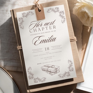 May include: A wedding invitation with the text "Her next CHAPTER CELEBRATE OUR BRIDE TO BE Emilia" in elegant script. The invitation is on a light-coloured paper with floral accents and the date "18 OCTOBER 2025". It is tied with twine and a small tag.