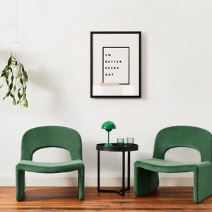 Pu&ograve; includere: Scena d'interni moderna con due sedie verde smeraldo con schienale arrotondato e design ritagliato. Un tavolino nero con una lampada verde e due bicchieri. Una stampa incorniciata con la scritta "1% BETTER EVERY DAY".
