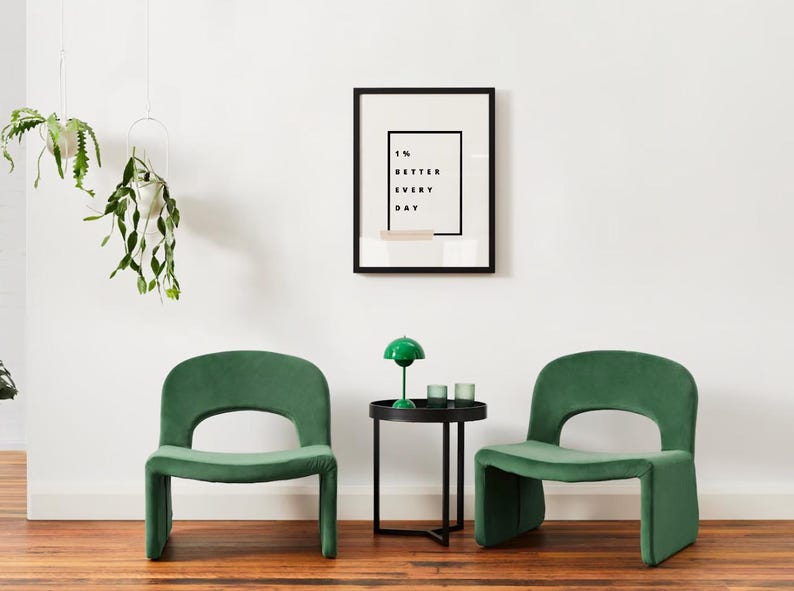 Pu&ograve; includere: Scena d'interni moderna con due sedie verde smeraldo con schienale arrotondato e design ritagliato. Un tavolino nero con una lampada verde e due bicchieri. Una stampa incorniciata con la scritta "1% BETTER EVERY DAY".
