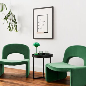 Pu&ograve; includere: Due poltrone in velluto verde smeraldo con schienale arrotondato e design ritagliato. Un tavolino nero con una lampada verde e due bicchieri si trova tra le sedie. Una stampa incorniciata &egrave; appesa alla parete bianca con la scritta "1% BETTER EVERY DAY."