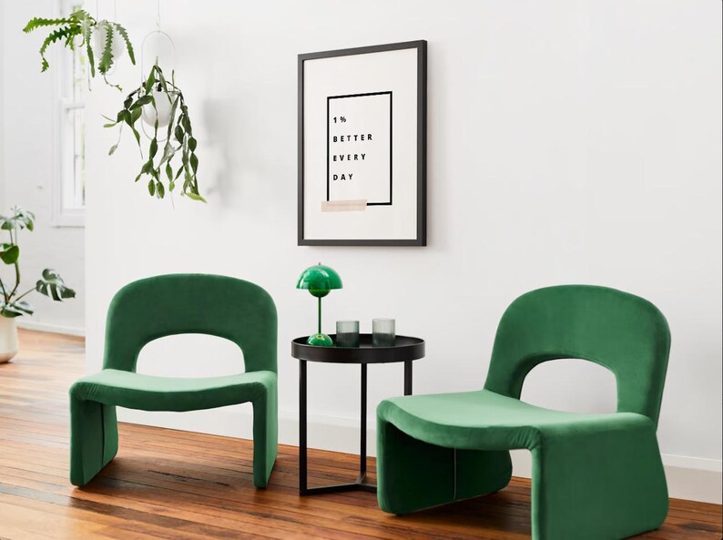 Pu&ograve; includere: Due poltrone in velluto verde smeraldo con schienale arrotondato e design ritagliato. Un tavolino nero con una lampada verde e due bicchieri si trova tra le sedie. Una stampa incorniciata &egrave; appesa alla parete bianca con la scritta "1% BETTER EVERY DAY."