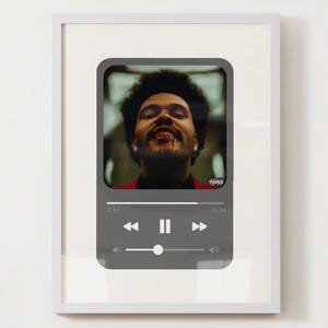 May include: Framed art print featuring a portrait of a man with dark curly hair, set within a gray music player interface. The image has a red and black color scheme. The frame is white.