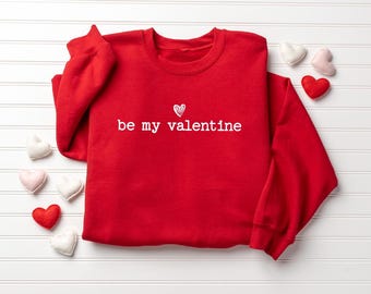 May include: A red sweatshirt with the phrase "be my valentine" in white script, accompanied by a small heart. Decorative felt hearts in red, pink, and white are scattered around the sweatshirt, creating a Valentine's Day theme.