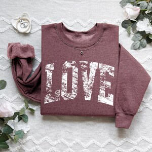 A burgundy sweatshirt with the word "LOVE" printed in white text is displayed on a white surface, surrounded by floral elements and a small white object.