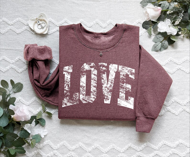 A burgundy sweatshirt with the word "LOVE" printed in white text is displayed on a white surface, surrounded by floral elements and a small white object.