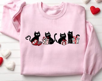 May include: A light pink sweatshirt featuring a graphic of four black cats with red and white gift boxes. The cats have heart-shaped tails. The gift boxes have red bows and heart designs. The sweatshirt is a crew neck style.
