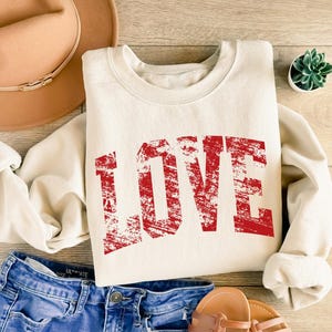 A casual outfit consisting of a white sweatshirt with the word "LOVE" printed in red, a pair of blue jeans, and a tan hat is laid out on a wooden surface.