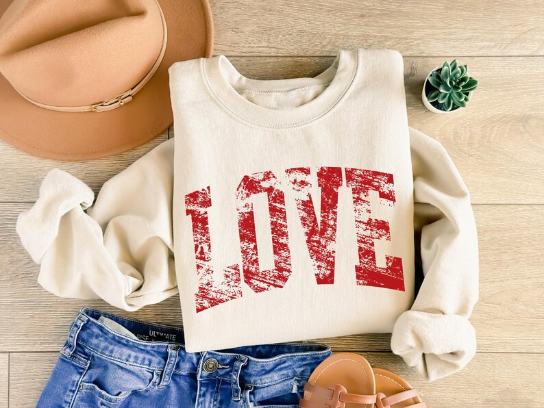 A casual outfit consisting of a white sweatshirt with the word "LOVE" printed in red, a pair of blue jeans, and a tan hat is laid out on a wooden surface.