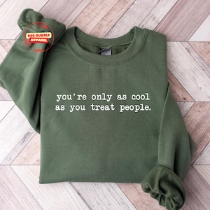 May include: An olive green sweatshirt with the text "you're only as cool as you treat people" in white. The sweatshirt is folded on a light wood surface. The Red Bubble Apparel logo is visible.
