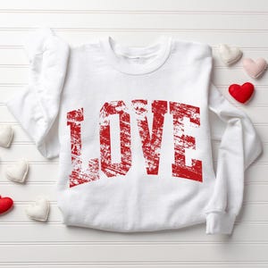 A white long-sleeved shirt with the word "LOVE" printed in red text, along with a few small heart-shaped objects surrounding it.
