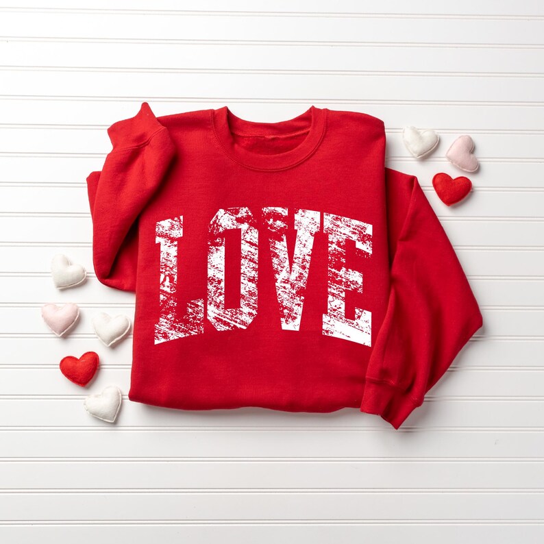 A red sweatshirt with the word "LOVE" printed in white text is displayed on a white background, surrounded by small white and pink heart-shaped objects.