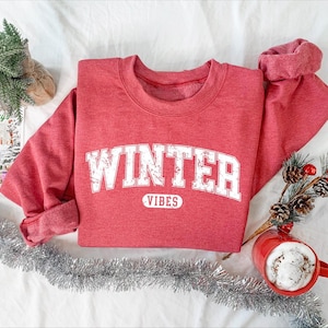 May include: A red sweatshirt with the words "WINTER VIBES" in white block letters. The sweatshirt is folded on a white surface, with a sprig of pine and a red mug filled with a white beverage.