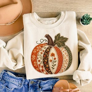 May include: A cream-colored crewneck sweatshirt featuring a large, central pumpkin graphic. The pumpkin is composed of various patchwork-style fabric patterns in shades of orange, brown, green, and white, with a dark brown stem and green leaf.