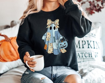 May include: Black sweatshirt featuring a patchwork ghost design with a bow and a pumpkin-shaped treat bag. The ghost is in shades of blue, beige, and brown. The sweatshirt is a comfortable, casual style, perfect for Halloween.