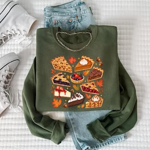 May include: An olive green crewneck sweatshirt featuring a graphic print of various pie slices, including apple, pumpkin, berry, cherry, and chocolate, decorated with autumn leaves and acorns, paired with blue denim jeans and white high-top sneakers.