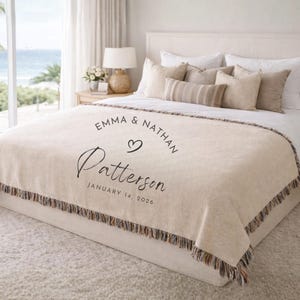 May include: A beige woven throw blanket with fringe edges, featuring the printed text EMMA & NATHAN, a heart icon, the name Patterson, and the date JANUARY 14, 2026. The blanket is draped over a bed with white pillows and a white headboard.