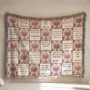 May include: A woven throw blanket featuring a grid pattern of alternating cream-colored squares with black religious text and squares with a dark red floral damask print. The blanket has a fringed edge with multi-colored threads along the sides.