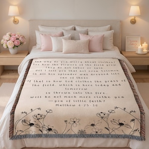 May include: A cream-colored woven throw blanket featuring black text from Matthew 6:25-30 and a black floral line art border at the bottom. The blanket has a fringed edge and is draped over a bed with white bedding and several pink and white pillows.