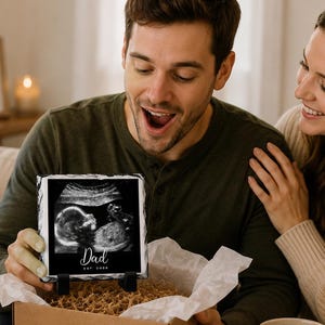 May include: A black and white ultrasound image on a slate tile, displayed on a stand, with the word "Dad" and the year "2000". The tile is in a gift box with shredded paper. A card reads "You're going to be a Dad!" with baby booties.