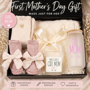 May include: A gift box for a new mother, containing a cream-colored blanket, a pair of pink baby socks with bows, a candle, and a glass with the text "GIRL MOM". The box is black and has the text "First Mother's Day Gift".