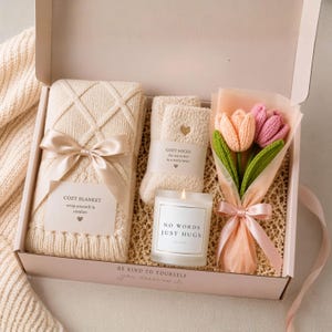 Overhead view of an open gift box with a folded cream knit blanket, fuzzy socks, a candle labeled NO WORDS JUST HUGS, and wrapped crocheted tulips.