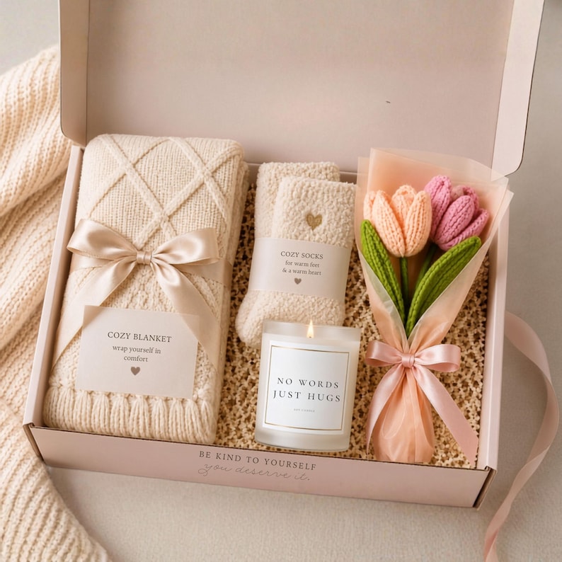 Overhead view of an open gift box with a folded cream knit blanket, fuzzy socks, a candle labeled NO WORDS JUST HUGS, and wrapped crocheted tulips.
