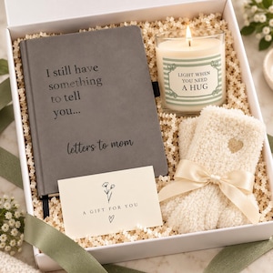 May include: A white gift box containing a gray journal with the text I still have something to tell you... letters to mom, a glass candle with a label reading LIGHT WHEN YOU NEED A HUG, cream fuzzy socks with a gold heart, and a card saying A GIFT FOR YOU.