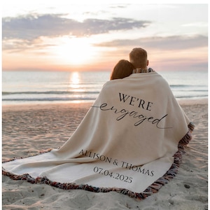 May include: A cream-colored blanket with the words "WE'RE engaged" in black script. Below the text are the names "ALLISON & THOMAS" and the date "07.04.2025". The blanket has a fringed edge with multiple colors. The blanket is on a beach.