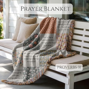 May include: A woven throw blanket featuring a patchwork design of floral patterns in muted earth tones and white text excerpts from Proverbs 31. The blanket has a fringed edge and is draped over a white bench. The text reads PRAYER BLANKET and PROVERBS 31.