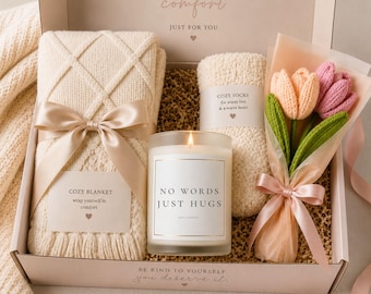 May include: A gift box containing a cream-colored knit blanket with a satin ribbon, a pair of fuzzy socks labeled 'COZY SOCKS', a lit candle with a label reading 'NO WORDS JUST HUGS', and a bouquet of three knitted tulips wrapped in pink paper.