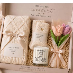 May include: A gift box containing a cream-colored cable knit blanket with a satin ribbon, a pair of fuzzy cream socks with a gold heart, a glass candle labeled "Smells like rest and recovery," and a bouquet of crocheted pink and purple tulips.