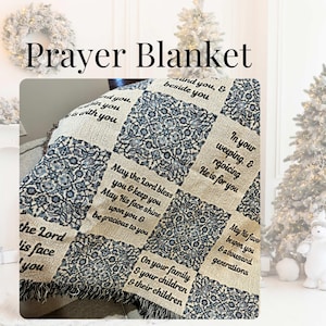 May include: A woven throw blanket featuring a patchwork design of cream-colored squares with black printed religious text and alternating blue and white intricate floral patterned squares, finished with a fringed edge along the bottom border.