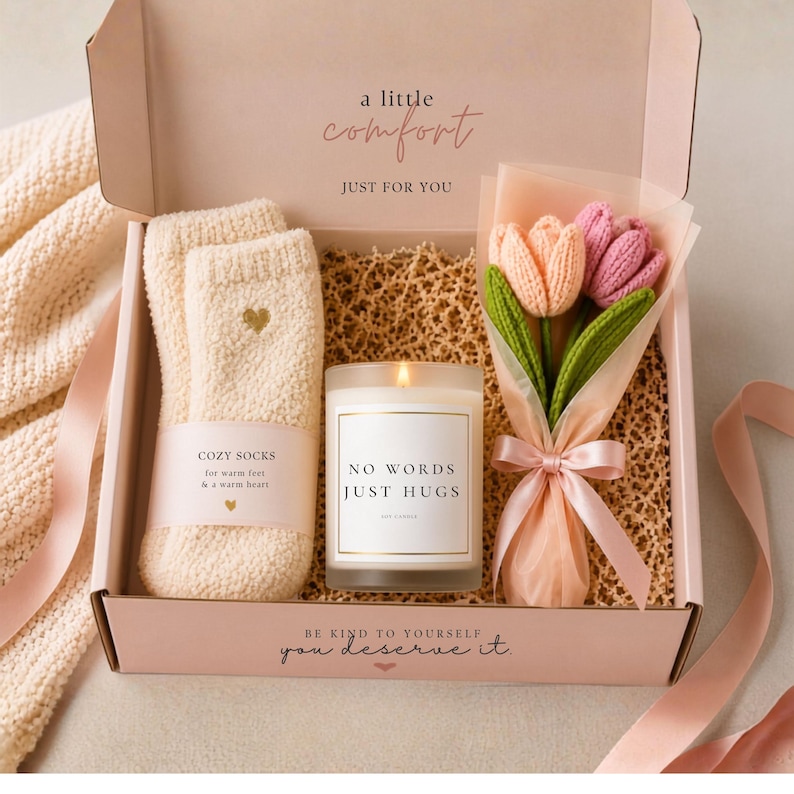 Open pink gift box containing fuzzy socks, a glass candle with NO WORDS JUST HUGS label, and a small bouquet of crocheted tulips on a crinkle paper bed.