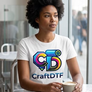 May include: A white t-shirt featuring a colorful logo with the text "CraftDTF". The logo is a stylized combination of letters and shapes in shades of blue, yellow, and pink. The person is sitting at a table with a cup of coffee.