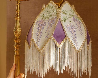 May include: A gold-colored floor lamp with a floral design. The lamp shade is white with purple and green floral embroidery, trimmed with gold and white beaded fringe. The lamp is held by a person's hand.