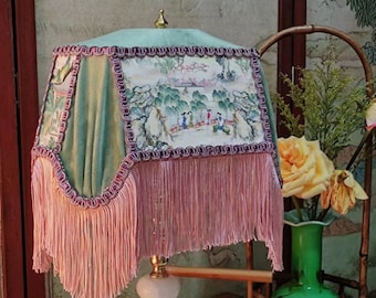 Può includere: Un paralume verde e bianco con frange rosa e una base dorata. Il paralume presenta una scena di paesaggio cinese tradizionale con figure in primo piano. La lampada è appoggiata su un tavolo con un libro e alcuni fiori secchi.