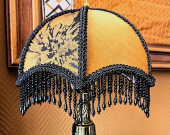May include: A gold table lamp with a black and gold patterned lampshade. The lampshade has a beaded fringe.