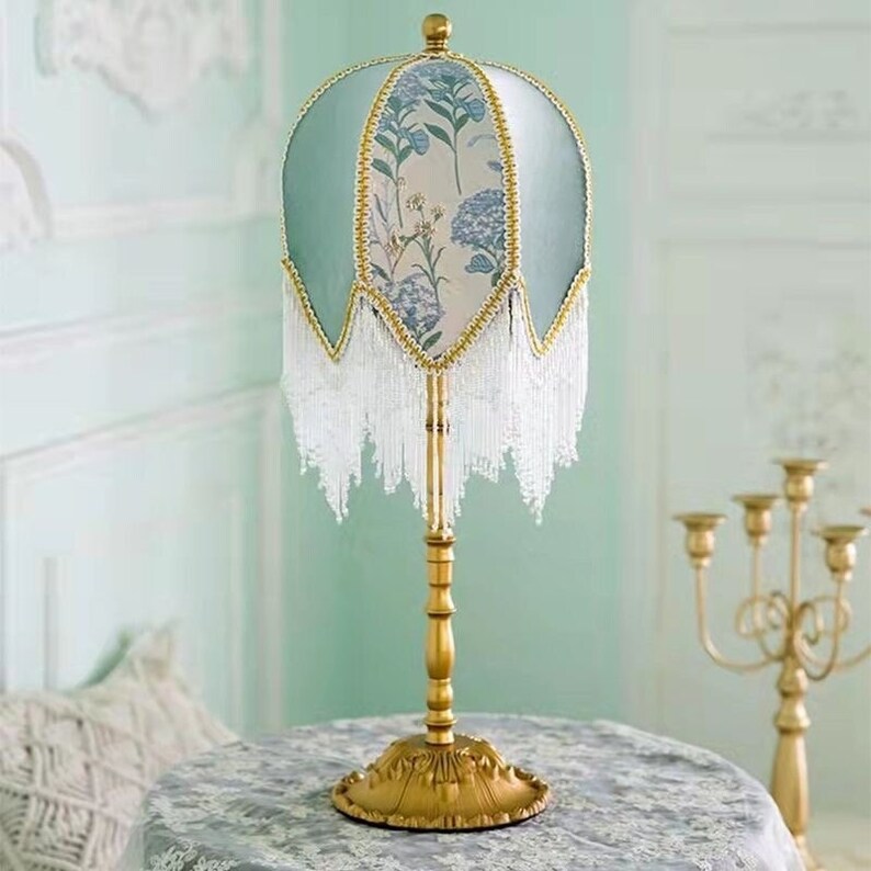 Puede incluir: Una l&aacute;mpara de mesa dorada con una pantalla de l&aacute;mpara con estampado floral blanco y azul. La pantalla tiene una franja de cuentas blancas.