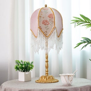 Puede incluir: Una l&aacute;mpara de mesa dorada con una pantalla rosa con estampado floral. La pantalla tiene un fleco de cuentas y la base de la l&aacute;mpara tiene un dise&ntilde;o decorativo.