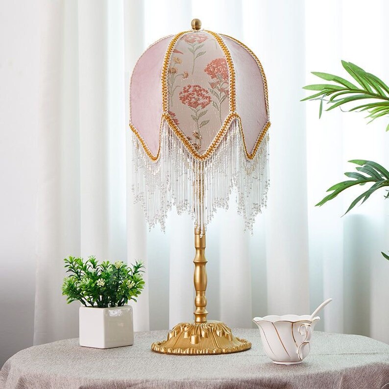 Puede incluir: Una l&aacute;mpara de mesa dorada con una pantalla rosa con estampado floral. La pantalla tiene un fleco de cuentas y la base de la l&aacute;mpara tiene un dise&ntilde;o decorativo.