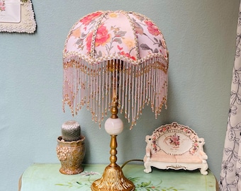 May include: A vintage table lamp with a floral patterned lampshade in pink and red. The lamp has a gold-coloured base and stem, with a white marble-like orb. The shade is adorned with beaded fringe.