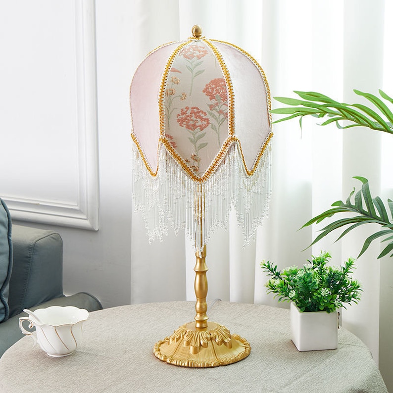 Puede incluir: Una l&aacute;mpara de mesa dorada con una pantalla de l&aacute;mpara de estampado floral rosa y blanco. La pantalla tiene una franja de cuentas blancas. La l&aacute;mpara est&aacute; sobre una mesa blanca con una taza y un platillo blancos y una planta verde en una maceta blanca.