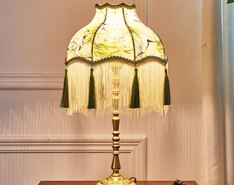May include: A gold table lamp with a white and green patterned lampshade. The lampshade has a fringe trim and features a floral design with birds.