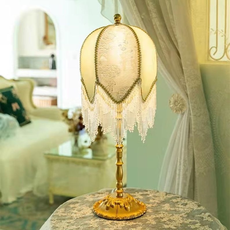 Puede incluir: Una l&aacute;mpara de mesa dorada con una pantalla de tela blanca y dorada con flecos de cuentas. La pantalla tiene un patr&oacute;n floral.