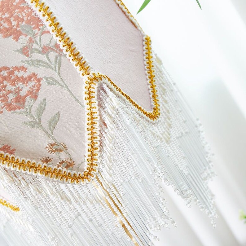 Puede incluir: Una pantalla de l&aacute;mpara rosa con un patr&oacute;n floral y flecos de cuentas blancas. La pantalla de la l&aacute;mpara tiene un borde dorado.
