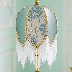 Puede incluir: Una pantalla de l&aacute;mpara azul claro con un patr&oacute;n floral y flecos blancos. La pantalla de la l&aacute;mpara est&aacute; hecha de tela y tiene un ribete dorado.