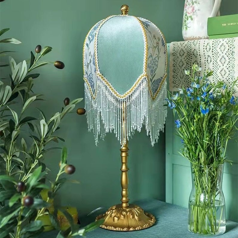 Puede incluir: Una pantalla de l&aacute;mpara verde y blanca con una base dorada y ribete de flecos. La pantalla tiene un patr&oacute;n floral y est&aacute; sentada sobre una mesa.